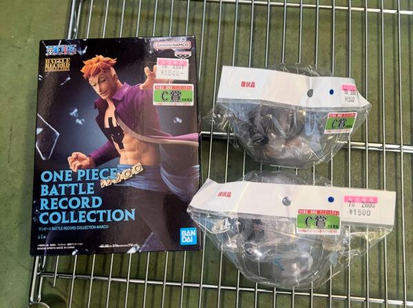 Img_e0702 - Articulos Japan (post Box Japan) Boxed One Piece Battle Record Collection figure with an orange-haired character on the cover, placed on a metal shopping cart next to two sealed prizes with price stickers Articulos Japan (Post Box Japan)