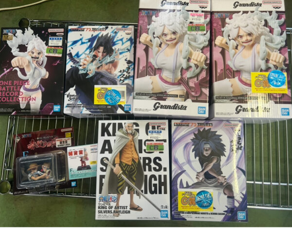 Img_e0709 - Articulos Japan (post Box Japan) Assortment of anime figure boxes on a metal shelf, featuring Grandista statues of Luffy and other characters with price stickers visible. Articulos Japan (Post Box Japan)