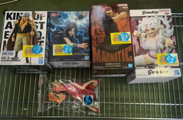 Img_e0711 - Articulos Japan (post Box Japan) Row of anime action-figure boxes on a metal rack, with a loose packaged figure in front. Articulos Japan (Post Box Japan)