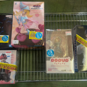 Boxed anime figures arranged on a metal wire rack, including a pink-haired character figure in the center and a Pop Up Parade Nobara Kugisaki box in front. Other boxed figures surround them with yellow price stickers visible.