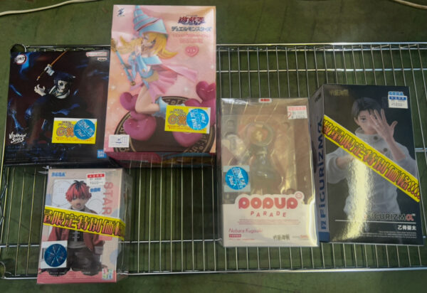 Img_e0713 - Articulos Japan (post Box Japan) Boxed anime figures arranged on a metal wire rack, including a pink-haired character figure in the center and a Pop Up Parade Nobara Kugisaki box in front. Other boxed figures surround them with yellow price stickers visible. Articulos Japan (Post Box Japan)