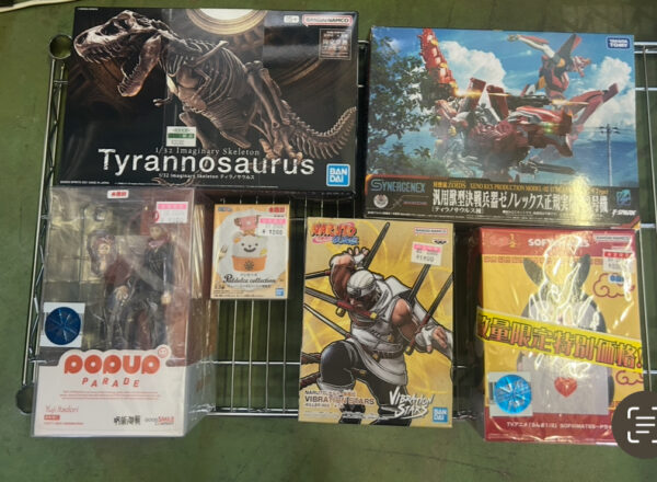 Img_e0722 - Articulos Japan (post Box Japan) Display of Japanese figure boxes on a wire rack: Tyrannosaurus skeleton figure, a blue transformer-style box, and anime figurines including Yuji Itadori (Pop Up Parade) and Naruto Vibration Stars set, with a yellow boxed item on the right. Articulos Japan (Post Box Japan)