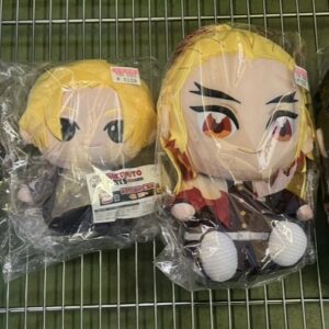 Four chibi anime plush dolls in plastic wrap on a metal rack, each wearing colorful outfits from left to right: teal checkered, two blond characters, and a dark-haired blue-eyed figure.