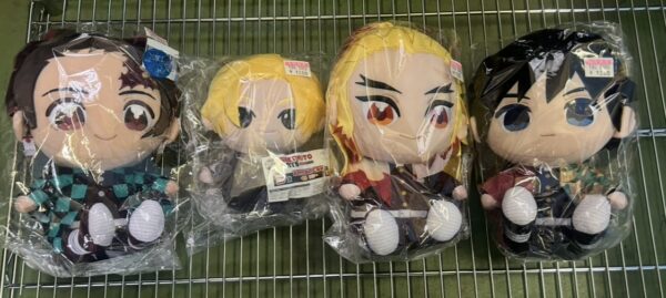 Img_e0724 - Articulos Japan (post Box Japan) Four chibi anime plush dolls in plastic wrap on a metal rack, each wearing colorful outfits from left to right: teal checkered, two blond characters, and a dark-haired blue-eyed figure. Articulos Japan (Post Box Japan)