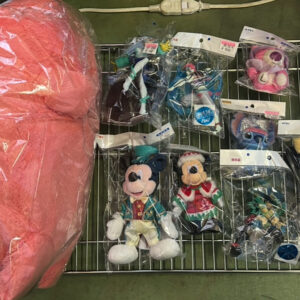 Wire rack on a workshop floor displays assorted sealed plush toys, including Disney characters, with a large pink stuffed item wrapped in plastic on the left.