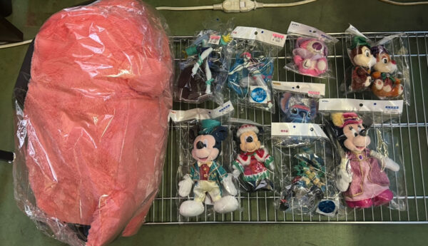 Img_e0728 - Articulos Japan (post Box Japan) Wire rack on a workshop floor displays assorted sealed plush toys, including Disney characters, with a large pink stuffed item wrapped in plastic on the left. Articulos Japan (Post Box Japan)