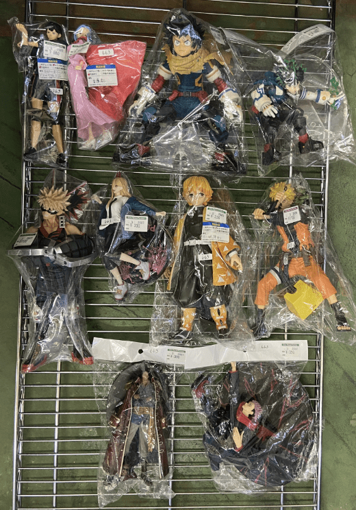 Ver Fotos Recientes 15 - Articulos Japan (post Box Japan) Assorted anime action figures in plastic bags on a metal display rack, arranged in rows on a green surface. Articulos Japan (Post Box Japan)