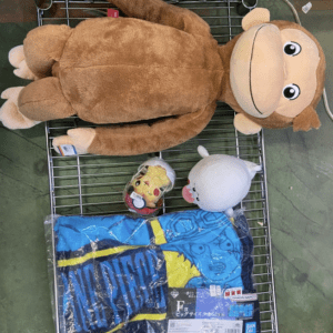 Large brown monkey plush lying in a shopping cart, with a white stuffed bunny, a small Pikachu figure in a capsule, and a blue folded garment with yellow accents visible in the cart.