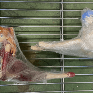 Two small plastic dolls wrapped in clear plastic cling to metal bars, one with red shoes and orange hair, the other showing a blue-streaked leg pose.