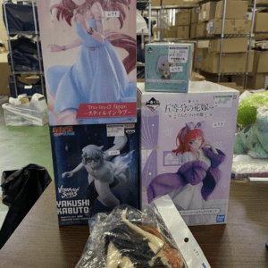 Boxes of anime figures on a table in a storage area; tall pink box with a girl in blue dress beside smaller blue-green and purple boxes, plus loose parts in plastic bag in front.
