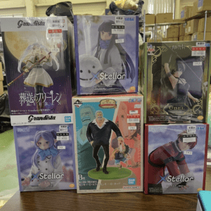 Stack of boxed anime figures on a store shelf featuring 'Grandista' and 'Stellar'シリーズ boxes with visible price stickers and SEGA/BAN/DA logos in a warehouse-like setting.