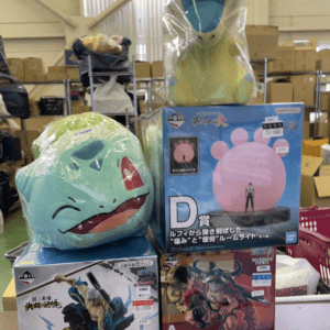Green frog-like plush resting on a blue toy box, with other boxed figures and a pink character image in a shop setting.