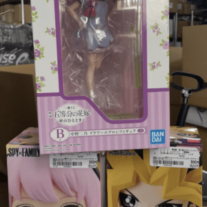 Stacked anime figure boxes in a store; top pink floral Ichiban Kuji window box with a red-haired figure, below are two Qposket boxes (pink-haired girl and blond boy).