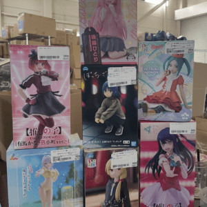 Row of boxed anime figures on a display table in a warehouse-style store, colorful character artwork visible on each box.