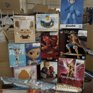 Stack of boxed anime figurines on a display shelf, including Mega Man Grandista and various One Piece, Pokémon, and Fate/strange fake figures with price stickers visible on the boxes.
