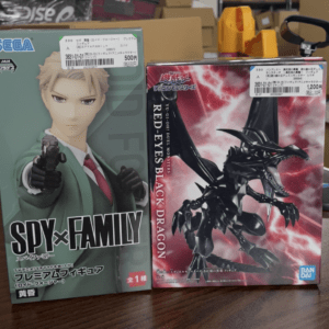 Two boxed action figures on a table: left shows a Spy×Family character in a green suit pointing a gun; right shows a Black Dragon from Yu-Gi-Oh! with a red background.