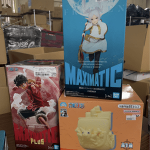 Boxes of anime figurines on a display table in a warehouse, with a tall blue Maximatic figure box featuring a white-haired character in the center.