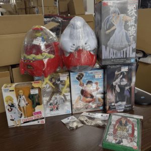 A display of assorted anime figure boxes and two large plastic-wrapped character figures on a table in a warehouse-like setting.