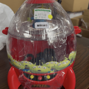 Red plastic prize dispenser with a clear dome lid, wrapped in plastic, decorated with green alien characters around the base.