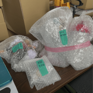 Bubble-wrapped bundles with green price tags labeled '642 ¥550' arranged on a wooden desk; a large wrap with pink tape, plus cardboard boxes and a yellow tool in the background.