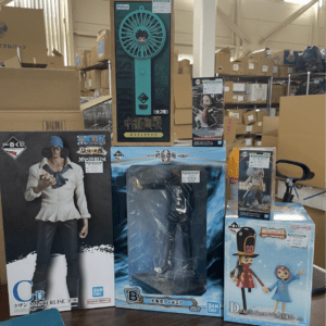 Display of anime figure boxes in a store, including a large blue standing figure box in the center with other boxed figures around it at the bottom shelf and cardboard boxes in the background.