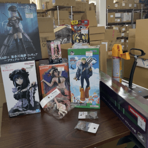 Boxed anime figures arranged on a wooden table in a storage room with shelves and cardboard boxes in the background.