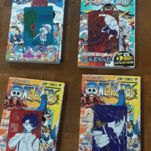 Four One Piece manga volumes laid on a wooden surface with colorful cover art and black-and-blue character cards attached to each book.