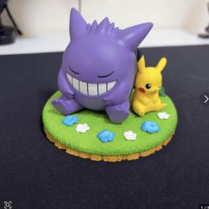 Purple Gengar toy with a wide grin sits on a grassy base beside a yellow Pikachu figure among blue and white flowers.