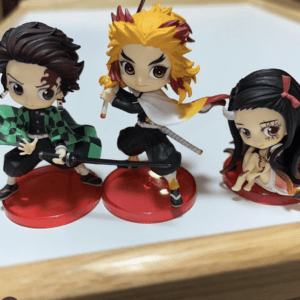 Three chibi Demon Slayer figures on red bases: Tanjiro with a green checkered cloak, wielding a sword; Rengoku in black with a white cape and yellow hair, in an action pose; Nezuko seated with a pink bow and stitched dress beside them.