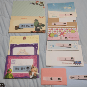 Colorful collection of decorative envelopes and cards laid out on a blue surface; left side features cartoon characters, right side has white envelopes labeled with numbers 1–6.