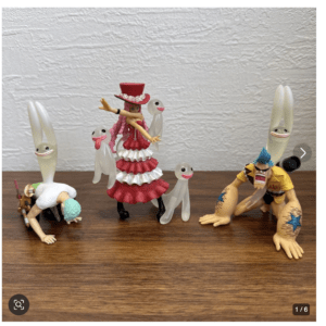 Group of anime-style figurines on a wooden table: a pink-dressed girl with a red hat surrounded by translucent ghost figures, plus a kneeling white-clad character and a muscular yellow-haired fighter on the right.