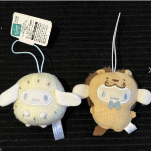 Two small pastel plush keychain toys with hanging loops on a dark surface: a greenish-yellow creature with white ears and a brown bear-like figure with a blue bow tie.