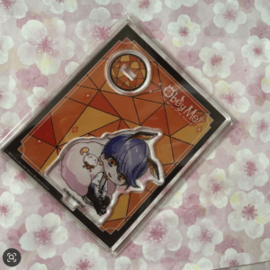 Merchandise packaging with a chibi anime-style character (purple hair) and a pink sheep figure against an orange geometric backdrop on a floral pink background.