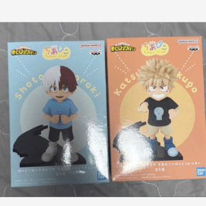 Two boxed chibi figures from My Hero Academia: Shoto Todoroki (blue box) and Katsuki Bakugo (orange box) on a gray quilted surface, each figure standing on a black base.