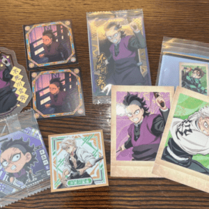 Assorted anime trading cards and mini prints on a wooden surface, featuring a male character in a purple outfit in various action poses, some in plastic sleeves.  Articulos Japan (Post Box Japan)