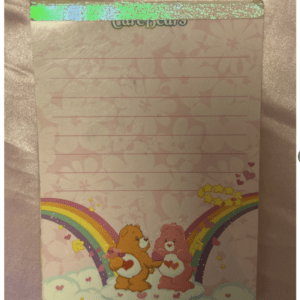Notebook with pink floral background, teal glitter tape at the top, and a Care Bears illustration of two bears on a rainbow at the bottom.