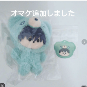 Turquoise plush toy with a bear hood in a plastic bag, with a small green badge beside it; white Japanese text overlays the image.
