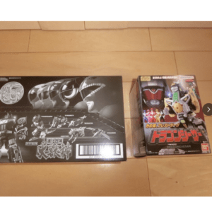 Two toy boxes on a wooden floor: a large black dinosaur-themed box left and a Dragon Caesar boxed figure right.