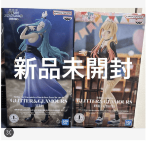 Two Bandai Namco Glitter & Glamours anime figure boxes side by side, showing a blue-haired figure on the left and a blonde figure on the right with bold white Japanese text overlay.