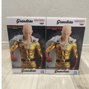 Two Grandista action figure boxes featuring Saitama from One Punch Man, gold suit and white cape, side by side on a wooden floor.