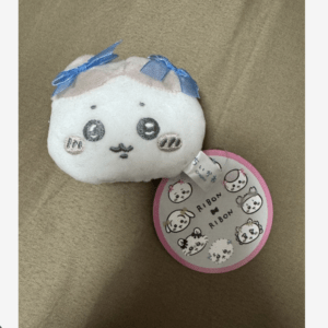 White plush stuffed toy with round face, blue ribbon bows on top, and a pink circular tag featuring tiny animal illustrations.