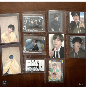 Collection of K-pop photo cards stored in clear plastic sleeves on a wooden surface, featuring several individual members in various poses and outfits.