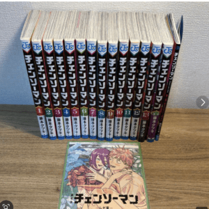 Row of Japanese manga volumes lined upright on a wooden table, spines with bold white Japanese text and color accents; a colorful front cover shows two characters in an embrace laid on the table in front of the row.