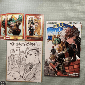 My Hero Academia collection: three red-bordered hero cards, two character cards, a black-and-white group sketch, and a large color manga volume featuring Deku and Bakugo on a gray surface.