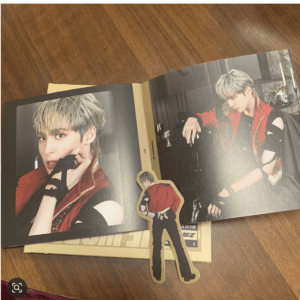 Open fashion magazine spread on a wooden table featuring a male model with bleached hair in a red and black outfit, plus a cardboard cutout figure in front.