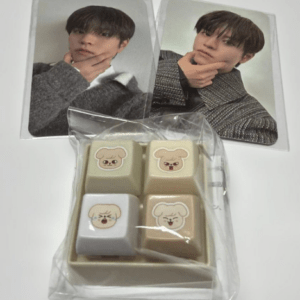 Gift set with four decorative keycaps featuring cute animal faces in a plastic bag, placed beside two photo cards of a young man in a gray sweater.  Articulos Japan (Post Box Japan)