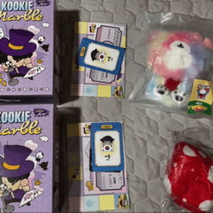 Two purple Kookie Marble toy boxes with a card set and small items on a quilted bedspread; a rainbow plush in a plastic bag nearby.