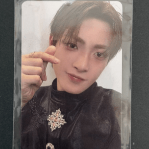 Portrait of a young man with light brown hair in a black outfit, wearing a jeweled brooch, making a finger heart gesture.
