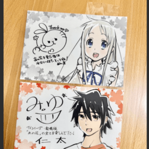 Two illustrated cards on a wooden table: top card shows a white-haired girl with a blue ribbon beside a bunny doodle, bottom card features a black-haired boy in an orange shirt with cherry-blossom borders and handwritten text.