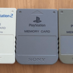 Three PS2 memory cards on a wooden table: a white 8MB MagicGate card on the left and two gray Sony memory cards to the right.